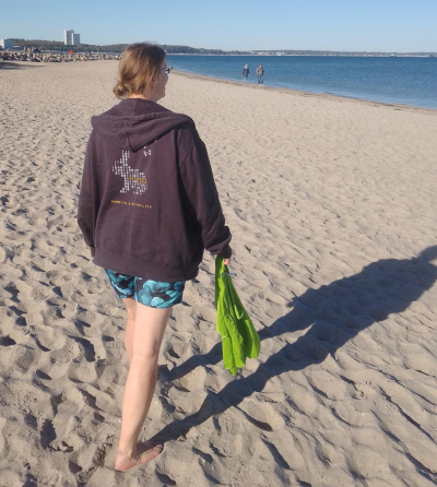 Das Foto zeigt mich von hinten am Strand. Der Strand ist fast leer. Ich trage eine Badehose mit Quallen drauf und eine Hoodie-Jacke mit einem Easterhack-Aufdruck. Die Beine sind bis Mitte Oberschenkel nackt, die HAltung ist gerade, in der rechten Hand baumelt ein kleines, grünes Handtuch. Die Haare sind in einem Knoten zusammengebunden, aber einige sind lose.