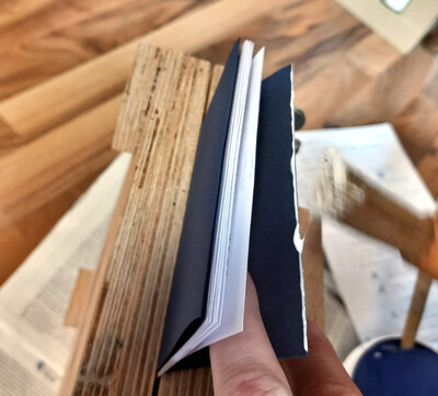 Finger halten das Vorlagepapier des in die Presse geklemmten Buches ab, um die obere Innenkante mit einer einen Milli Meter dicken Leimschicht einzupinseln. Es handelt sich um die plattgepfalzte Kante.