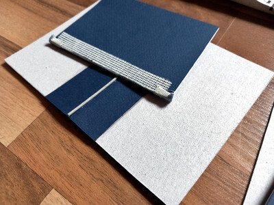 Auf einer noch nicht eingeschlagenen Buchumschlagpappe liegt ein geleimter Buchblock mit Vorsatzpapier, an den nun auch oben und unten das Kapitalbändchen angeleimt ist. Es ist hellblau oder hellgrau, und insgesamt so 2-3 Millimeter lang.